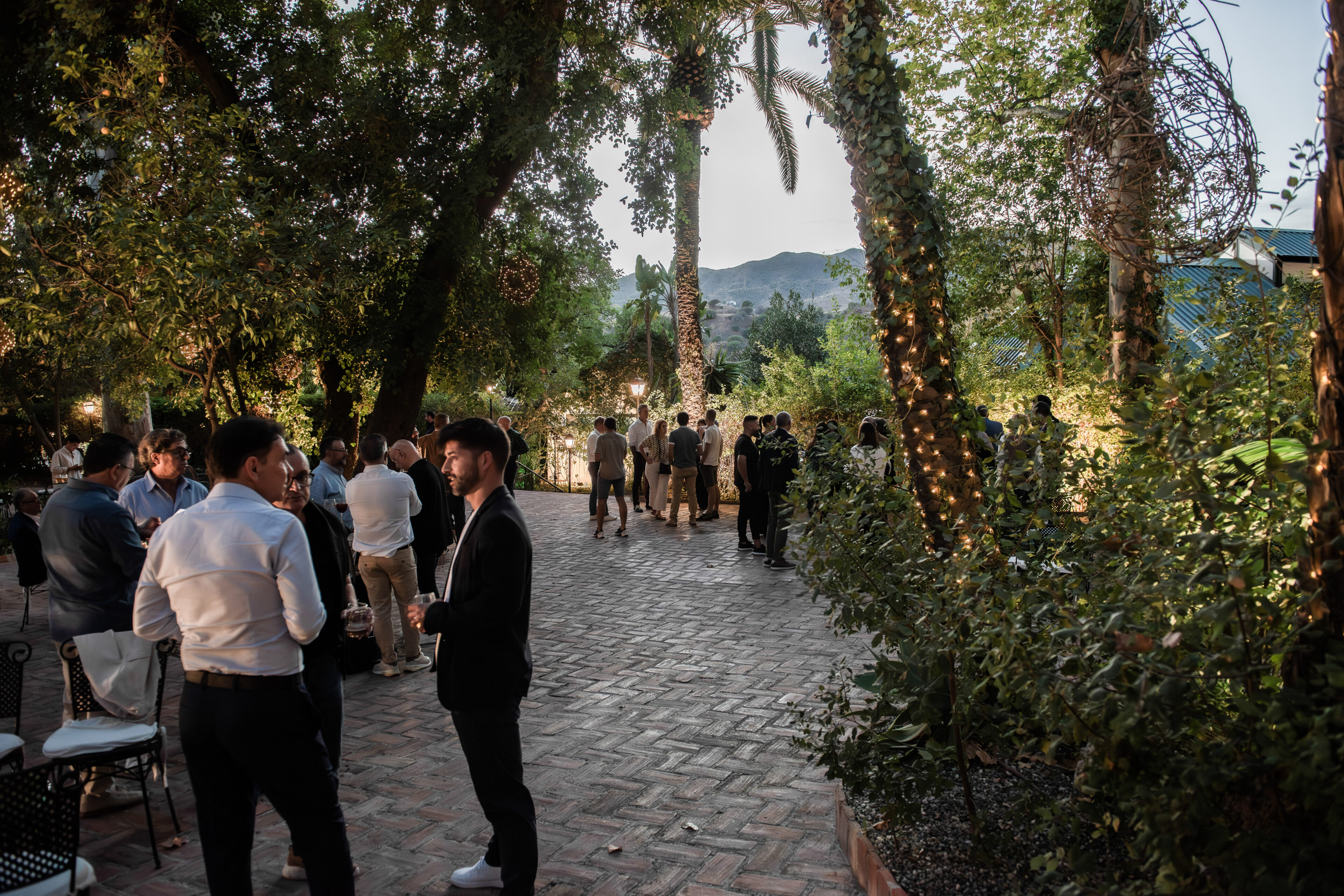 Grupo de personas en un jardín exterior con pavimento de ladrillo y árboles iluminados, ambiente sofisticado de evento social nocturno. NEXO Küchen Grupo de personas en un jardín exterior con pavimento de ladrillo y árboles iluminados, ambiente sofisticado de evento social nocturno. NEXO Küchen