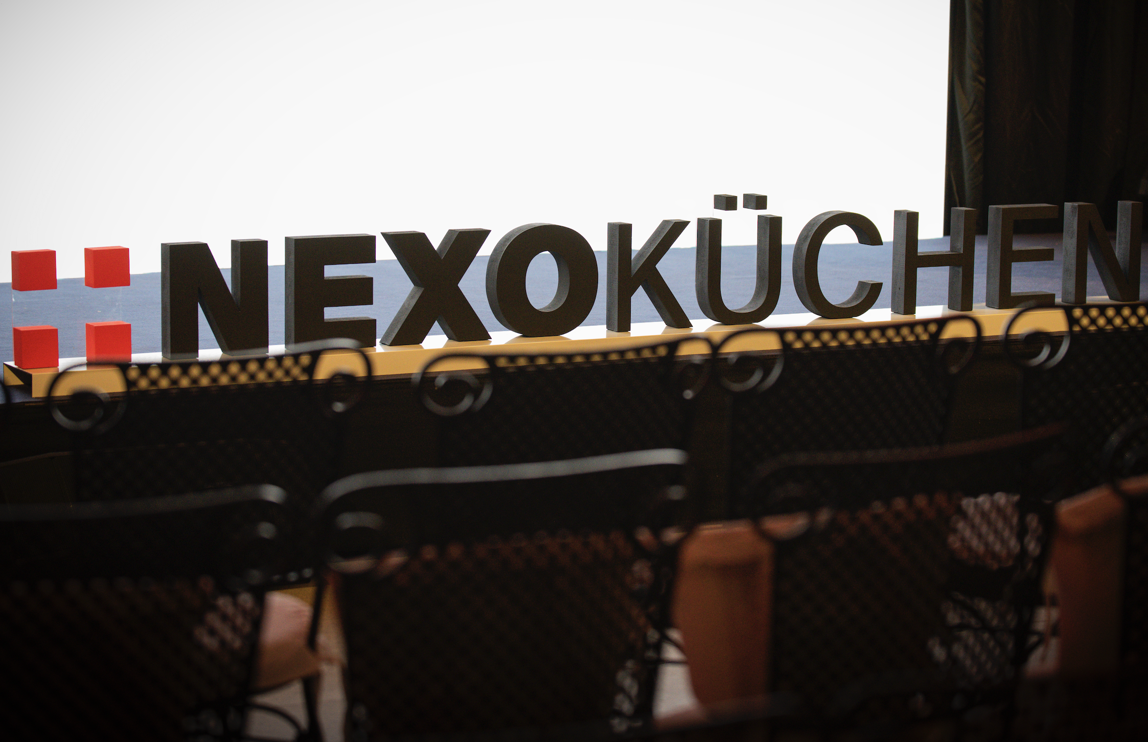 NEXO Küchen gran cartel de letras negras NEXO KÜCHEN en un escenario, barandilla amarilla y sillas negras al frente. NEXO Küchen gran cartel de letras negras NEXO KÜCHEN en un escenario, barandilla amarilla y sillas negras al frente.